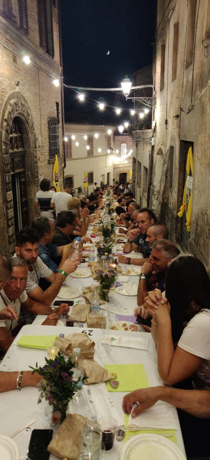 La tavolata della CenaLonga gremita di commensali, di sera, sotto le luci appese tra le case del corso di Amandola